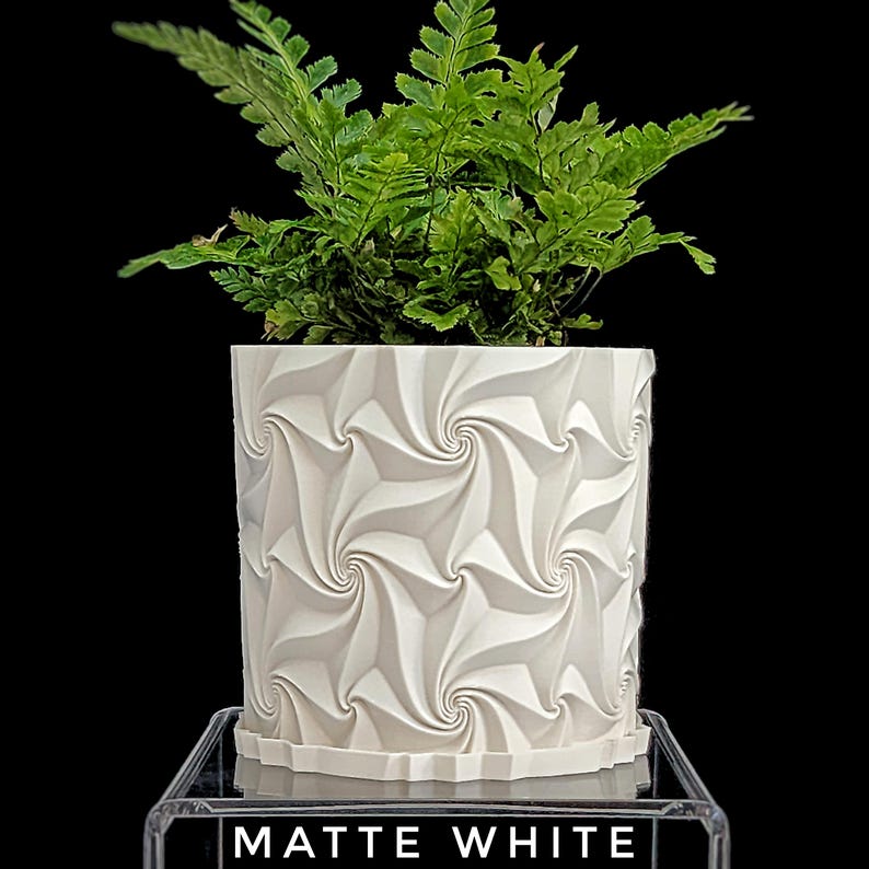 May include: A matte white cylindrical planter featuring a repeating geometric spiral pattern on its exterior surface. A vibrant green fern plant is potted inside the container. The base of the planter rests on a clear stand with the text MATTE WHITE.