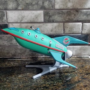 Pu&ograve; includere: Un modello di razzo di colore verde acqua e bianco, tratto dalla serie televisiva degli anni '60 "Perduti nello spazio". Il razzo &egrave; su un piedistallo bianco e ha le parole "Jupiter 2" sul lato.
