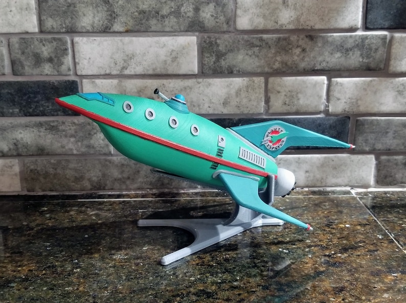 Pu&ograve; includere: Un modello di razzo di colore verde acqua e bianco, tratto dalla serie televisiva degli anni '60 "Perduti nello spazio". Il razzo &egrave; su un piedistallo bianco e ha le parole "Jupiter 2" sul lato.
