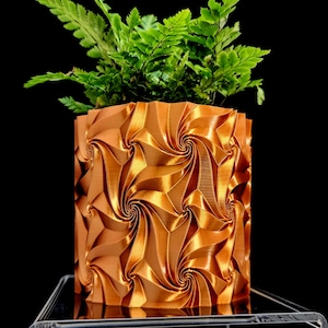 May include: A metallic copper-colored cylindrical planter featuring a repeating geometric pattern of swirling, vortex-like indentations. A vibrant green fern plant with delicate, feathery fronds grows out of the top of the textured container.