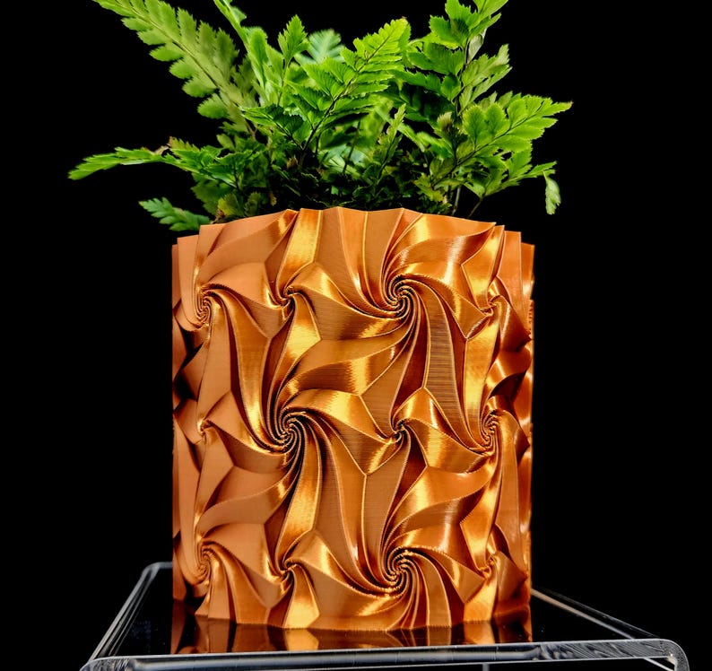 May include: A metallic copper-colored cylindrical planter featuring a repeating geometric pattern of swirling, vortex-like indentations. A vibrant green fern plant with delicate, feathery fronds grows out of the top of the textured container.