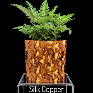 May include: A vibrant green fern plant potted in a metallic copper-colored cylindrical planter featuring a repeating geometric spiral pattern. The base of the planter rests on a clear acrylic stand with the text Silk Copper printed on the front.