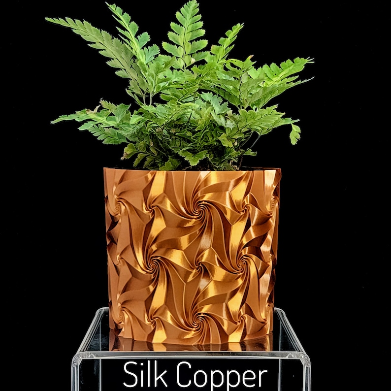 May include: A vibrant green fern plant potted in a metallic copper-colored cylindrical planter featuring a repeating geometric spiral pattern. The base of the planter rests on a clear acrylic stand with the text Silk Copper printed on the front.