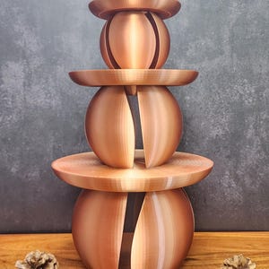 Op de afbeelding: Een decoratief object in koperkleur met drie niveaus. Het ontwerp heeft twee bolvormige vormen met verticale uitsparingen, gescheiden door platte, cirkelvormige platforms. Het bovenste element is een kleinere, bolvormige vorm. Twee dennenappels aan de basis.