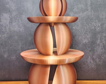 May include: A three-tiered, copper-colored decorative object. The design features two spherical shapes with vertical cutouts, separated by flat, circular platforms. The top element is a smaller, spherical shape. Two pinecones are at the base.