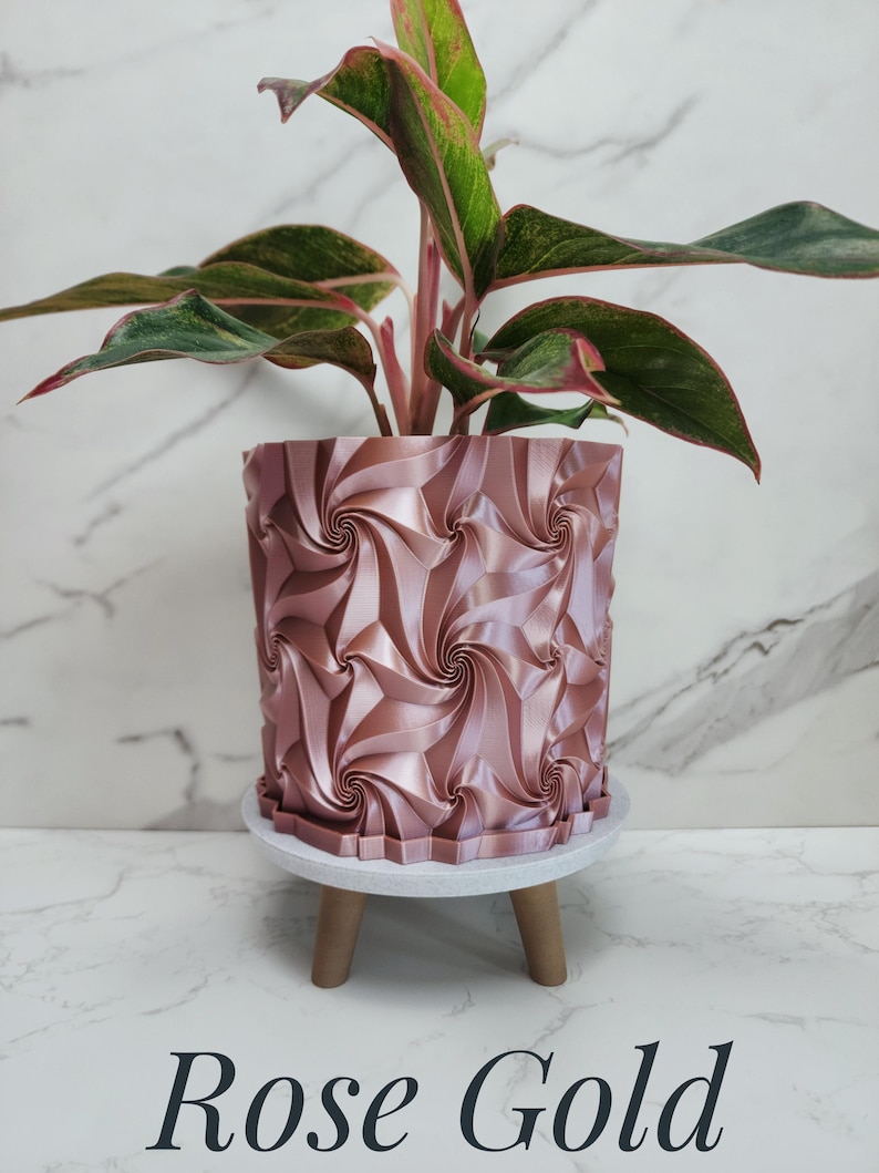 May include: A rose gold cylindrical plant pot with a repeating geometric spiral pattern. The pot sits on a white circular base with three short wooden legs. A plant with green and pink variegated leaves is planted inside the container.
