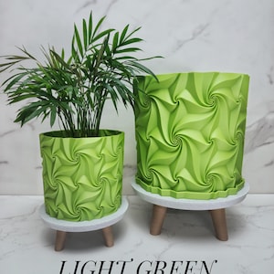 May include: Two light green cylindrical planters with a repeating geometric swirl pattern. The planters sit on small, round, light-colored stands with three short legs. A green plant is inside the smaller planter. The text LIGHT GREEN is below.
