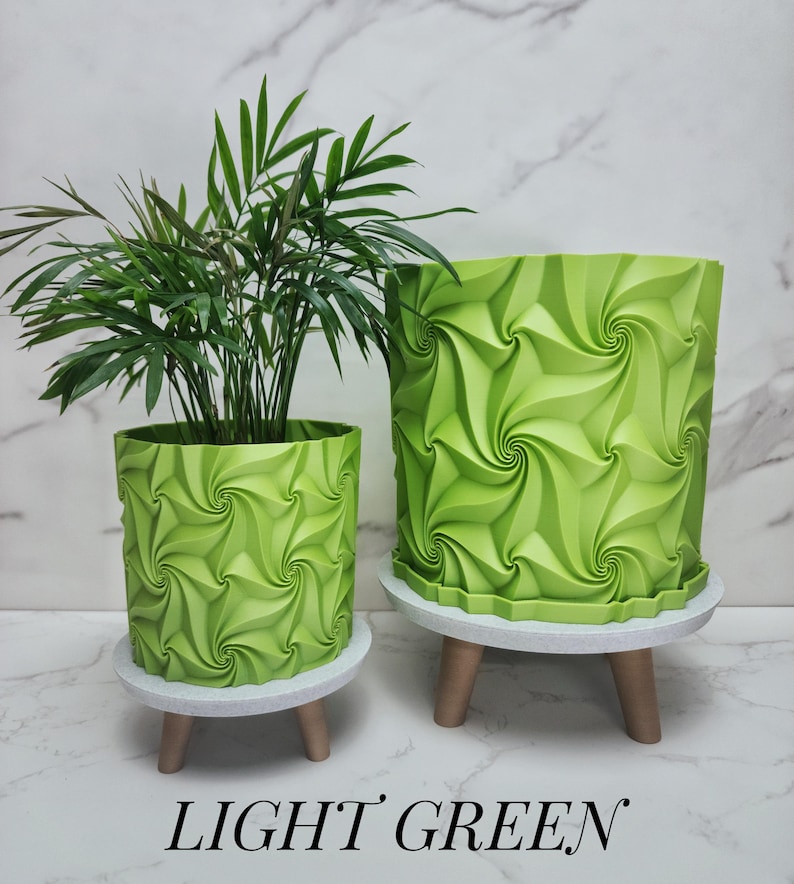 May include: Two light green cylindrical planters with a repeating geometric swirl pattern. The planters sit on small, round, light-colored stands with three short legs. A green plant is inside the smaller planter. The text LIGHT GREEN is below.