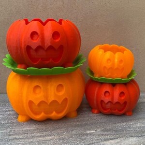 Peut inclure: Trois pots de fleurs en forme de citrouille orange et rouge avec des visages souriants. Le plus grand pot est empil&eacute; sur un pot plus petit, et un troisi&egrave;me pot encore plus petit est pos&eacute; sur la table &agrave; c&ocirc;t&eacute; d'eux.
