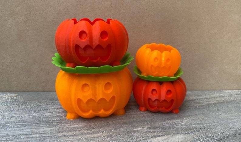 Peut inclure: Trois pots de fleurs en forme de citrouille orange et rouge avec des visages souriants. Le plus grand pot est empil&eacute; sur un pot plus petit, et un troisi&egrave;me pot encore plus petit est pos&eacute; sur la table &agrave; c&ocirc;t&eacute; d'eux.
