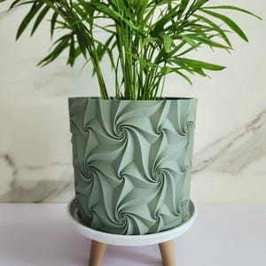 May include: A muted green cylindrical plant pot featuring a repeating geometric spiral pattern on its exterior. The pot rests on a white circular saucer supported by three short, light-colored wooden legs, containing a green leafy plant.