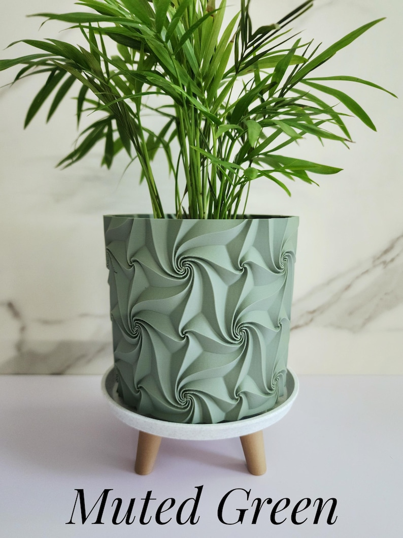 May include: A muted green cylindrical plant pot featuring a repeating geometric spiral pattern on its exterior. The pot rests on a white circular saucer supported by three short, light-colored wooden legs, containing a green leafy plant.