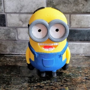 Pu&ograve; includere: Una statuina di un minion dei film Cattivissimo Me, stampata in 3D di colore giallo. Il minion indossa una salopette blu e ha un grande sorriso con denti bianchi. Il minion ha grandi occhiali rotondi grigi con occhi bianchi.