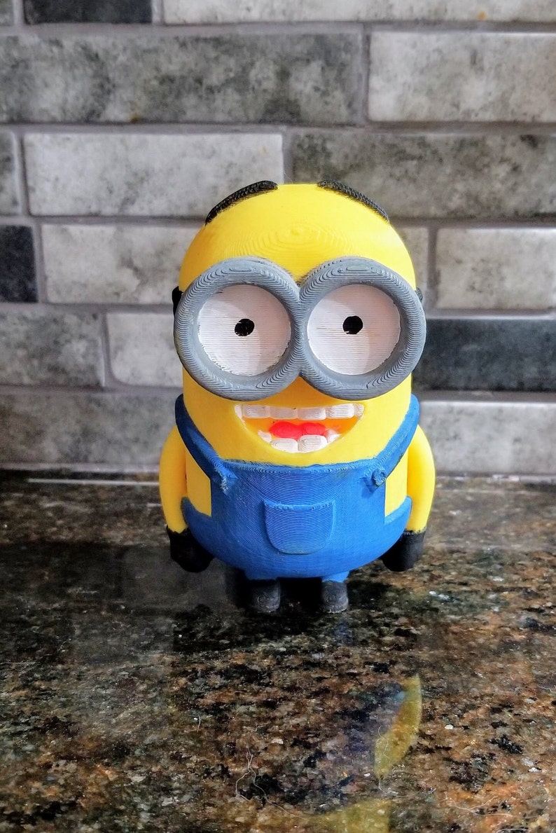 Pu&ograve; includere: Una statuina di un minion dei film Cattivissimo Me, stampata in 3D di colore giallo. Il minion indossa una salopette blu e ha un grande sorriso con denti bianchi. Il minion ha grandi occhiali rotondi grigi con occhi bianchi.