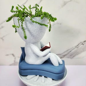 Pode incluir: Um vaso cinza-claro salpicado em forma de figura sentada num sofá azul a ler um pequeno livro. A cabeça da figura serve de vaso para uma planta verde pendente. A estrutura repousa sobre uma base circular cinza-claro com pernas.