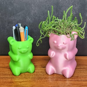 May include: Two green and pink gummy bear shaped planters. The green planter is holding pens and pencils. The pink planter has a small green plant growing out of it.