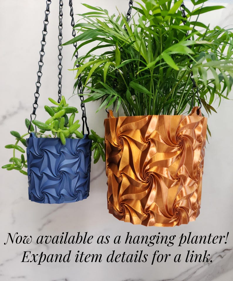 May include: Two hanging planters with a geometric spiral pattern suspended by black chains. The smaller planter is matte navy blue containing a green succulent, and the larger planter is metallic copper containing a green palm-like plant.
