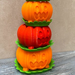 Peut inclure: Une pile de quatre pots en forme de citrouille orange et rouge avec des bases feuillues vertes. Chaque pot a un visage de citrouille sculpt&eacute;.