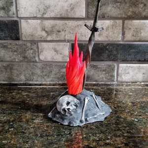 Pu&ograve; includere: Una statuetta stampata in 3D di un fal&ograve; con una spada che sporge dalla cima. Il fal&ograve; &egrave; fatto di plastica grigia e ha una fiamma rossa. C'&egrave; un teschio alla base del fal&ograve;.