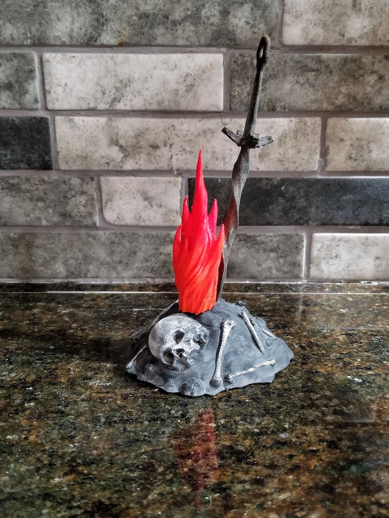 Pu&ograve; includere: Una statuetta stampata in 3D di un fal&ograve; con una spada che sporge dalla cima. Il fal&ograve; &egrave; fatto di plastica grigia e ha una fiamma rossa. C'&egrave; un teschio alla base del fal&ograve;.