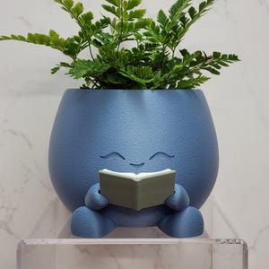 Może przedstawiać: Niebieska doniczka z uśmiechniętą twarzą, trzymająca małą zieloną książkę. Doniczka jest wypełniona zieloną paprocią. Doniczka stoi na przezroczystiej półce.