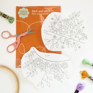 May include: Embroidery supplies including a package with "Stitch Happy" and "Stick and stitch" text, floral pattern transfers, pink scissors, embroidery floss, and a wooden hoop. The transfers feature floral designs.