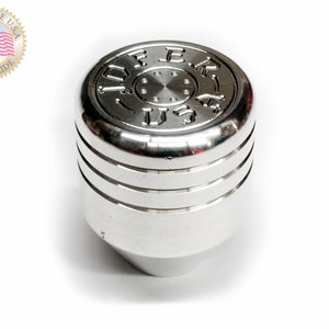 Puede incluir: Pomo de palanca de cambios plateado con una parte superior circular que presenta el texto "FEK U.S.".