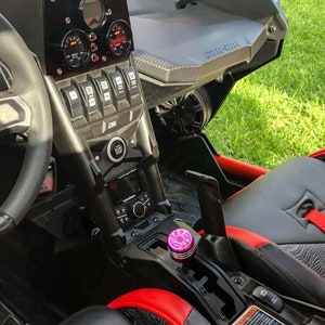 Puede incluir: El interior de un veh&iacute;culo todoterreno negro y rojo con un panel de instrumentos personalizado, un volante negro y una perilla de cambio p&uacute;rpura. El veh&iacute;culo tiene un logotipo de Can-Am en el tablero.