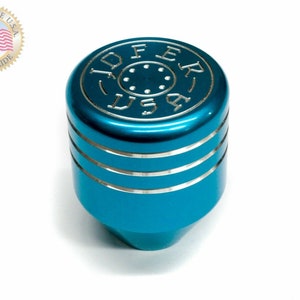 Puede incluir: Una palanca de cambios de color azul turquesa con una parte superior plateada y una inscripci&oacute;n plateada que dice "JOFER USA".