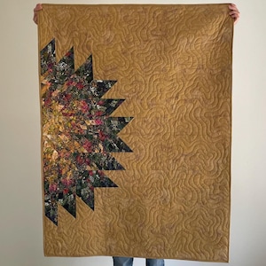 May include: A handmade quilt with a floral design on the left side, featuring a mix of red, green, and yellow hues. The right side of the quilt is a solid mustard color with a wavy pattern. The quilt is held up by a person.