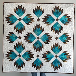 May include: A handmade quilt featuring a repeating pattern of turquoise and brown bear paw designs on a white background. The quilt is bordered with a brown fabric and has a quilted texture. The overall design is symmetrical and geometric.