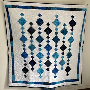 May include: A handmade quilt with a white background and a pattern of blue diamond shapes. The diamonds vary in shades of blue and are arranged in vertical rows. The quilt has a blue border and a black binding.