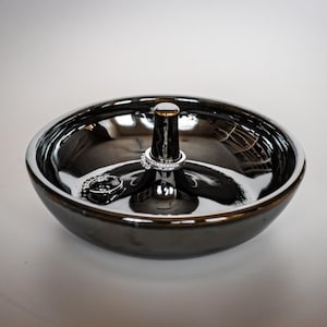 May include: A dark, metallic ring dish with a central post and a wide, shallow bowl. Two rings are displayed: one on the post and another in the bowl. The dish has a reflective surface, and the overall design is modern and minimalist.