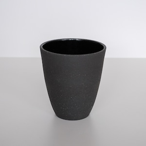 May include: A black ceramic cup with a textured surface and a slightly flared shape. The interior is a glossy black, contrasting with the matte exterior. The cup has a simple, modern design.