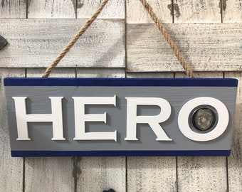May include: Wooden sign with the word "HERO" in white block letters. The sign is gray with a blue border and a rope for hanging. A police badge is embedded in the letter "O".