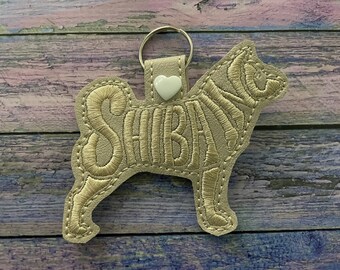 May include: A beige leather keychain shaped like a Shiba Inu dog with the word "SHIBA" embroidered in gold thread. A small white heart charm is attached to the key ring.