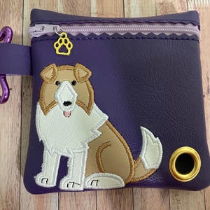 Op de afbeelding: Paarse ritszak met een hondenontwerp. De hond is een bruine en witte Collie met een witte borst en een bruine staart. De tas heeft een gouden metalen ring aan de zijkant.