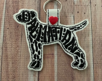 May include: Black and white embroidered dog silhouette keychain with a red heart charm. The dog is outlined in black and filled with a black and white pattern.