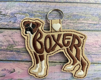 May include: A tan leather keychain with a brown embroidered boxer dog silhouette. The dog's name "BOXER" is embroidered in brown letters inside the silhouette. A white heart charm is attached to the key ring.
