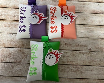 May include: Three zippered pouches with white fabric and embroidered text "Strike $$$." Each pouch has a different colored fabric on the back: purple, pink, and green. Each pouch has a small white bowling ball with red flames embroidered on it.