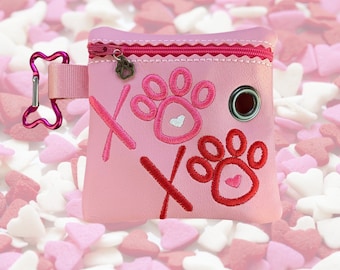 May include: Pink dog poop bag holder with a zipper closure and a paw print design. The bag holder has a clip for attaching to a leash. The bag holder has the text "XOXO" on it.