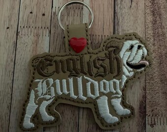 May include: Tan leather keychain in the shape of an English Bulldog. The keychain has the words "English Bulldog" embroidered in white and brown thread. A small red heart is attached to the top of the keychain.