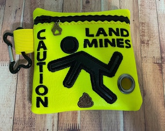May include: A bright yellow pouch with a black zipper and a black paw print charm. The pouch has black embroidered text that reads "CAUTION LAND MINES" and a black silhouette of a person tripping over a brown poop emoji.