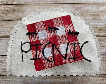 Op de afbeelding: Witte stof met een rood-wit geruit patroon en het woord "PICNIC" geborduurd in zwart. Er zijn vier zwarte mieren geborduurd op het geruite patroon.