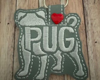 May include: A gray felt key chain with a white embroidered pug silhouette and the word "PUG" inside the silhouette. A red heart is above the word "PUG".