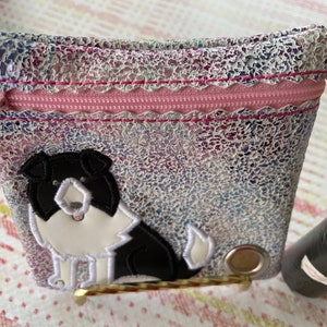 Op de afbeelding: Een klein, iriserend, met een rits te sluiten tasje met een roze rits en een zwart-witte border collie hond applique. Het tasje ligt op een roze en wit gepatroneerd oppervlak.