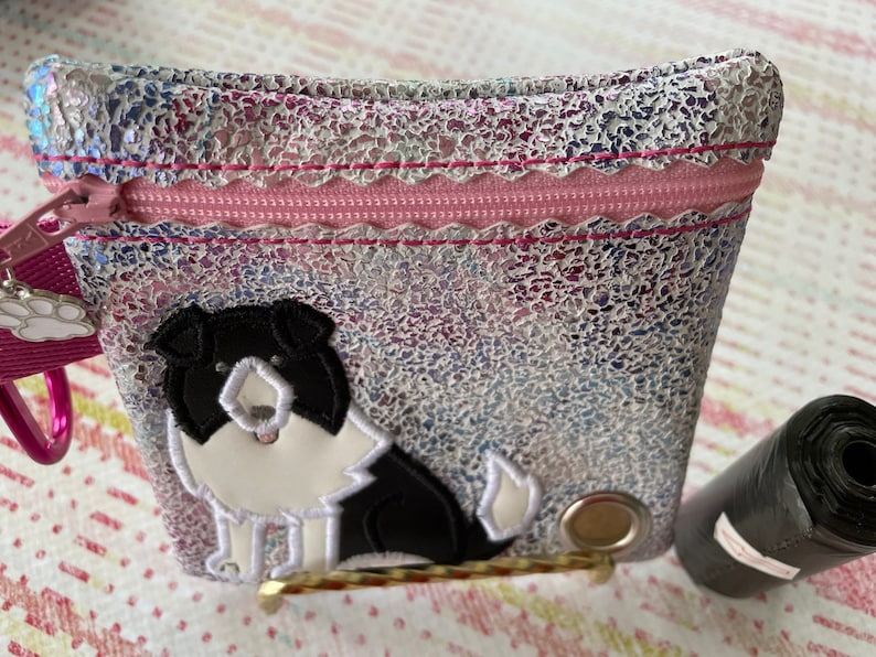 Op de afbeelding: Een klein, iriserend, met een rits te sluiten tasje met een roze rits en een zwart-witte border collie hond applique. Het tasje ligt op een roze en wit gepatroneerd oppervlak.
