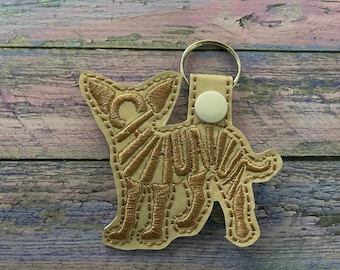 May include: A beige leather keychain with a gold embroidered chihuahua silhouette. The keychain has a white plastic ring and a silver metal key ring.