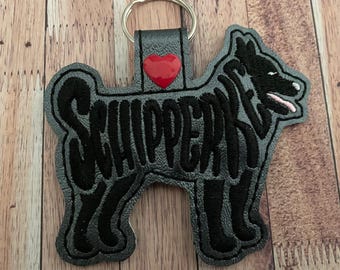 May include: A gray and black Schipperke dog-shaped keychain with a red heart accent. The word "SCHIPPERKE" is embroidered in black on the dog's body. The keychain has a silver metal ring attached to a black leather strap.