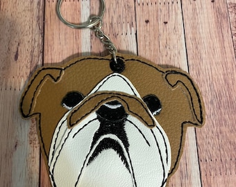 May include: A brown and white bulldog face keychain with black eyes and a black nose. The keychain is made of faux leather and has a silver metal ring.